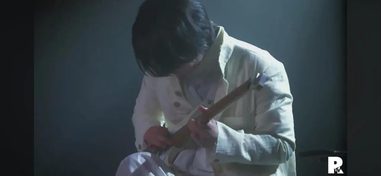 Ein Musiker in weißer Kleidung spielt eine Gitarre. Die Szene ist düster beleuchtet, was die Konzentration auf die Performance von Ichika Nito verstärkt.