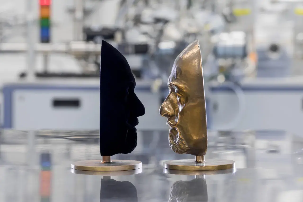Zwei Gesichtsmasken stehen sich gegenüber: eine aus samtigem, dunkelstem Schwarz, die andere aus glänzendem Bronze.