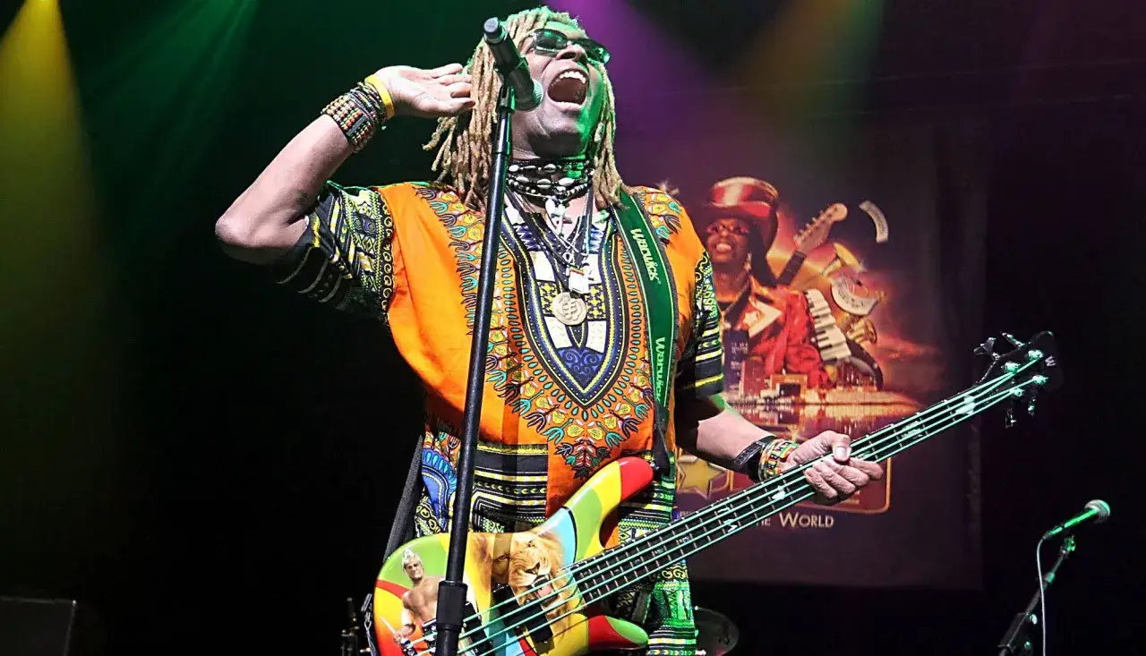 TM Stevens, mit Dreadlocks und Sonnenbrille, singt und spielt Bass auf einer bunten Gitarre.