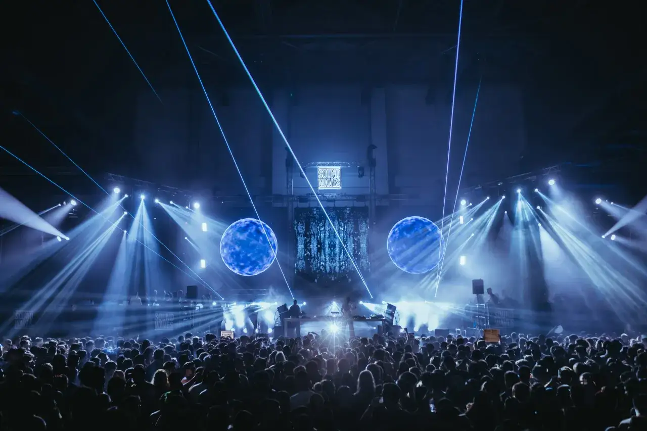 Eine riesige Menge tanzt im Takt von techno beats, w&auml;hrend blaue Laser und Scheinwerfer die B&uuml;hne in ein futuristisches Licht tauchen.