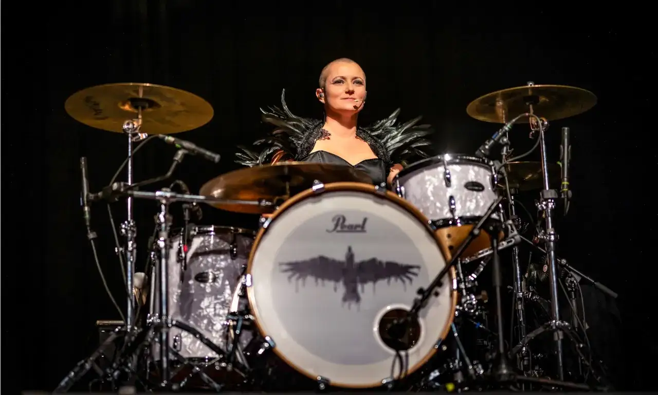 Katha Mia, eine Schlagzeugerin mit markantem Look, sitzt hinter ihrem Pearl-Schlagzeug. Ihr schwarzes Feder-Outfit und der Adler auf der Bassdrum sind beeindruckend.