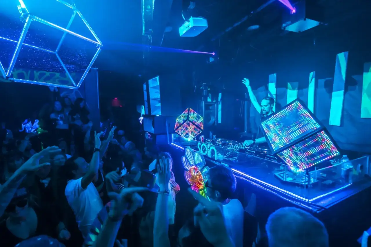 DJ steuert Lichtshow mit DMX-Software, w&auml;hrend Menge tanzt. Bunte W&uuml;rfel und Laserlicht schaffen pulsierende Atmosph&auml;re.