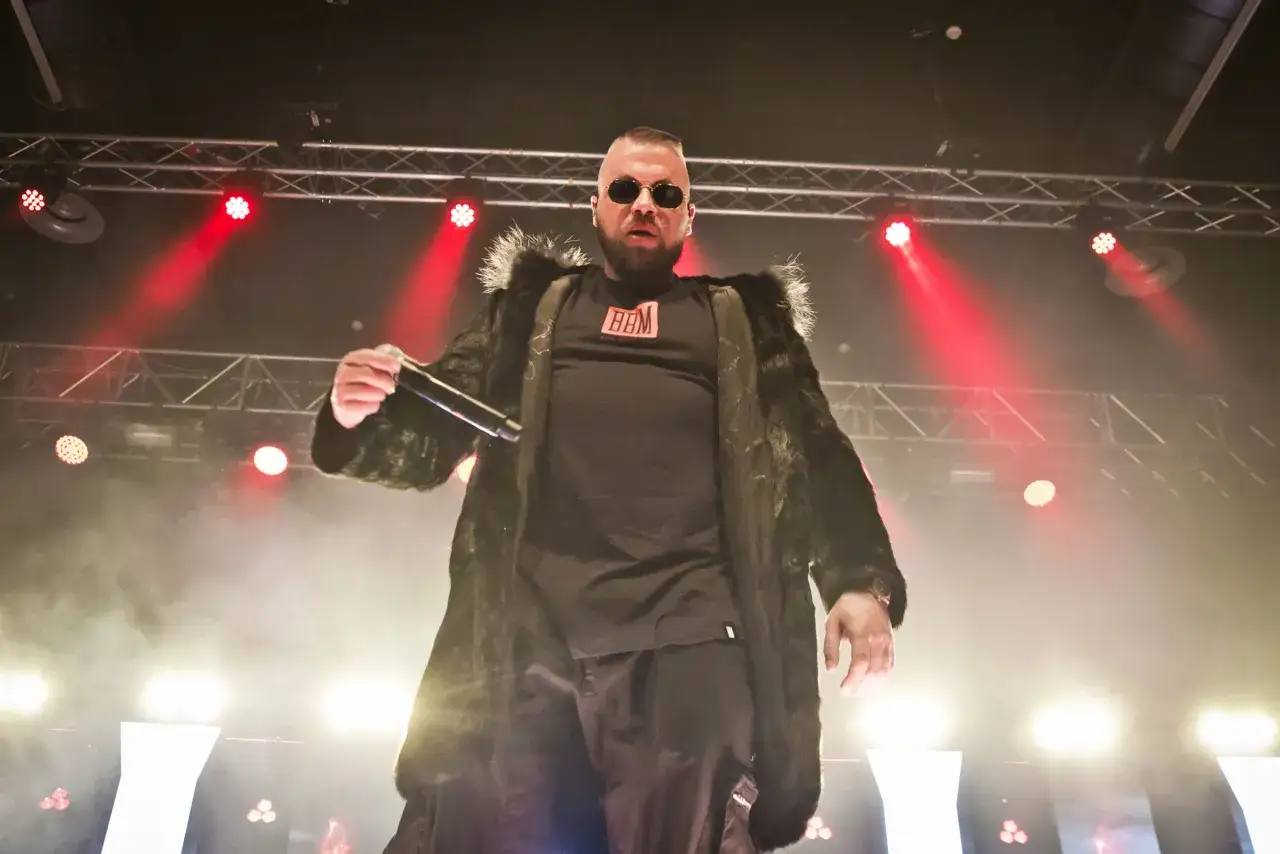 Rapper mit Sonnenbrille und Pelzmantel auf der B&uuml;hne, bereit, seine besten Kollegah Lines zu performen. Rote und wei&szlig;e Lichter im Hintergrund.