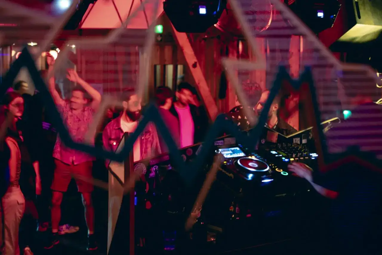 Menschen tanzen zu den dj sets, w&auml;hrend ein DJ am Mischpult steht. Rotes Licht und eine Grafik &uuml;berlagern die Szene.