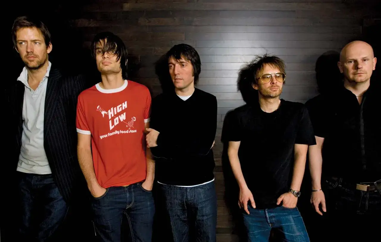 Die Mitglieder von Radiohead stehen nebeneinander vor einer dunklen Holzwand. Einer trägt ein rotes T-Shirt mit der Aufschrift