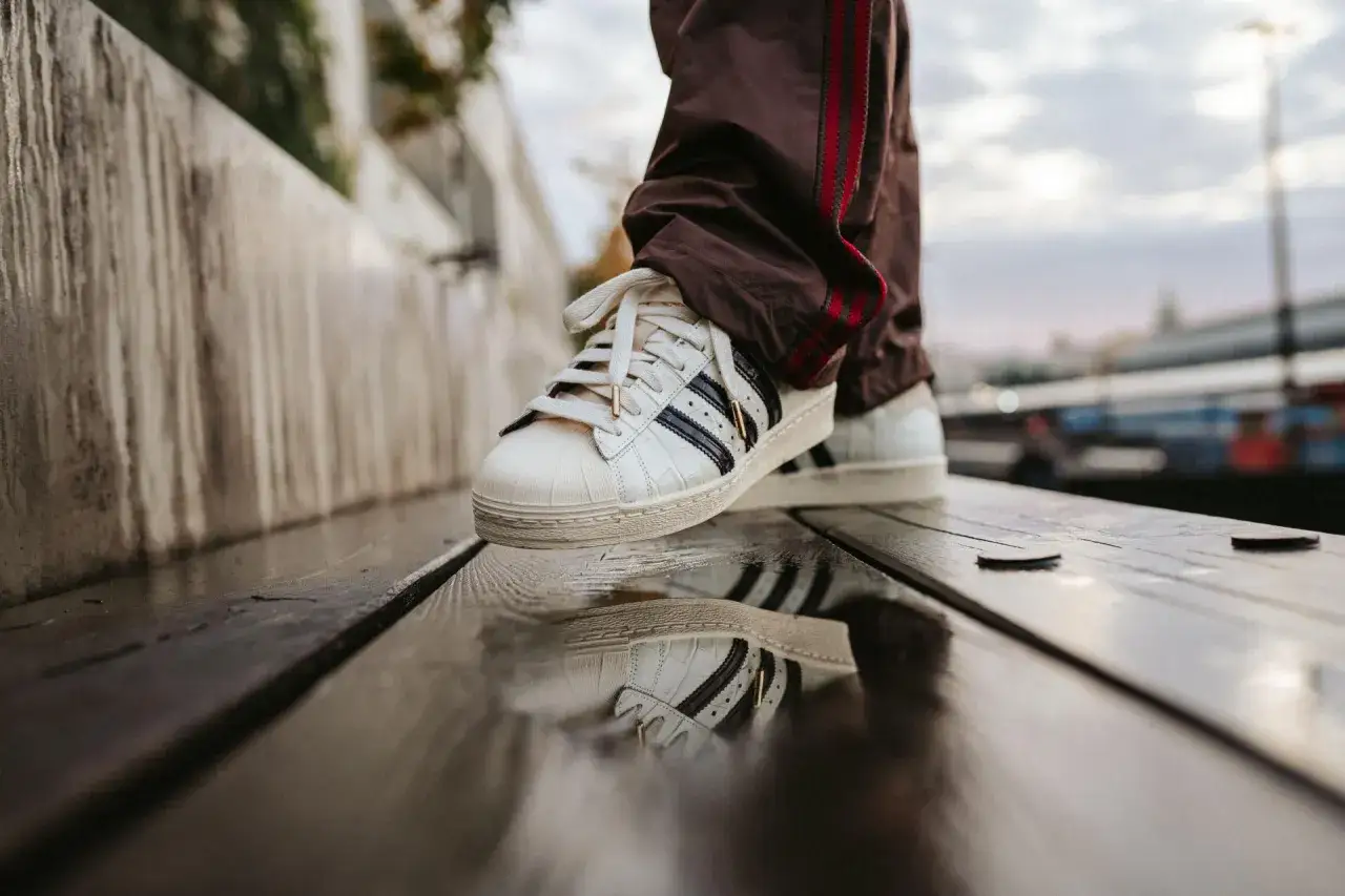 Weiße Adidas Superstar Sneaker mit Kroko-Prägung, Teil einer neuen Kollektion, spiegeln sich im nassen Holz.