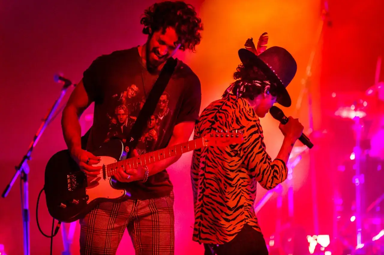 Alex Feder spielt Gitarre, während ein Sänger mit Hut und Tiger-Shirt auf der Bühne singt.