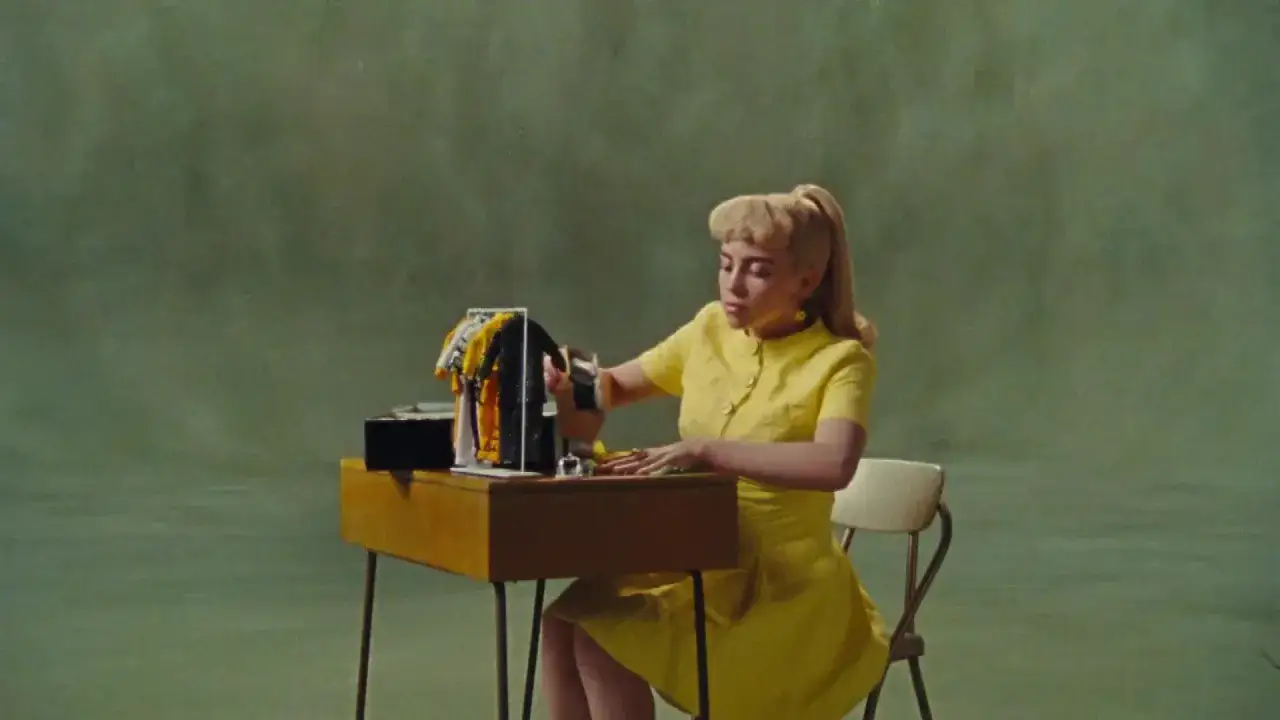 Billie Eilish in gelbem Kleid sitzt an einem Tisch und sortiert Kleiderpuppen. 
