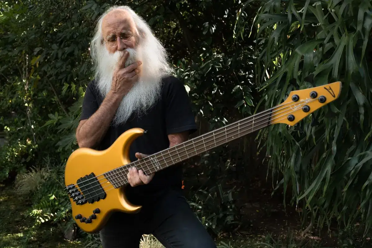 Lee Sklar, mit seinem ikonischen wei&szlig;en Bart, h&auml;lt eine goldene Bassgitarre in einem &uuml;ppigen Garten.