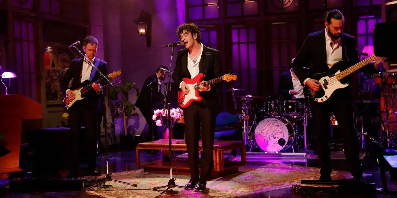 Die Mitglieder von The 1975 performen auf einer B&uuml;hne mit lila Beleuchtung. Ein Gitarrist spielt eine rote Gitarre, w&auml;hrend ein Bassist und ein weiterer Gitarrist daneben stehen.
