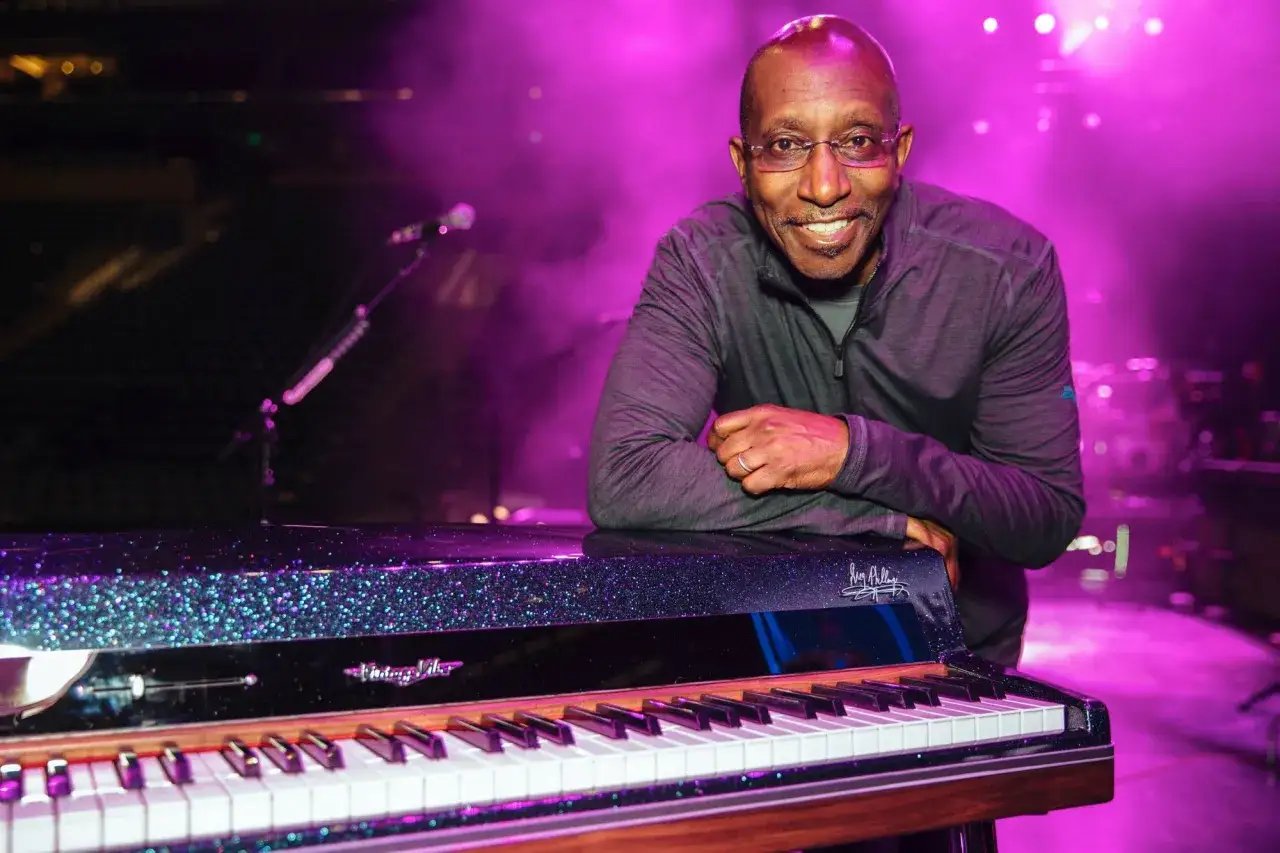 Greg Phillinganes l&auml;chelt neben einem glitzernden Klavier, im Hintergrund lila B&uuml;hnenlicht.