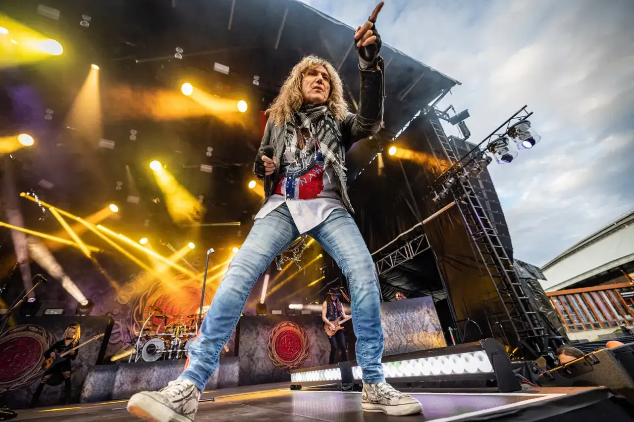 David Coverdale heute: Der Rock-Frontmann mit markanter Geste und voller Energie auf der B&uuml;hne, umgeben von Scheinwerfern und Bandkollegen.