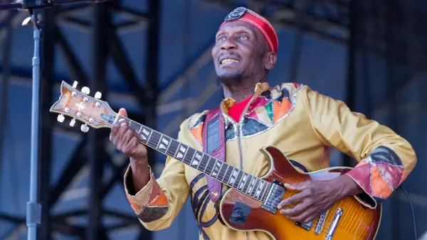 Ein verstorbener Musiker, eine Legende, spielt Gitarre auf der Bühne. Sein buntes Outfit und sein Ausdruck zeugen von seiner Leidenschaft.